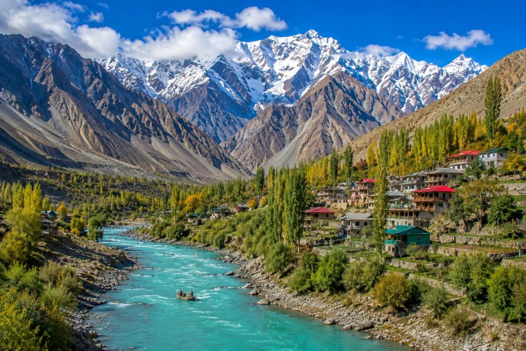 Panoramic view of Hunza Valley with turquoise river, colorful village houses, autumn trees, and snow-covered Karakoram mountains.