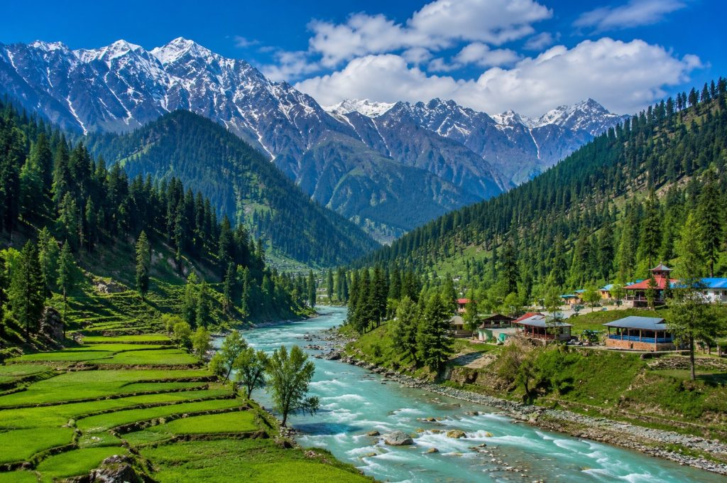 Beautiful landscape view of Swat Valley with flowing river, green mountains, pine trees, and snow-covered peaks under a blue sky.