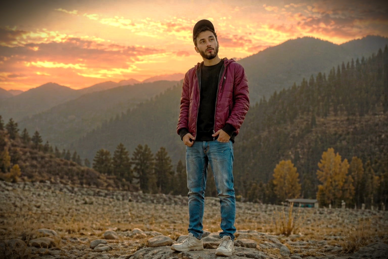 Young traveler standing on a rocky mountain landscape during sunset, surrounded by pine-covered hills and a colorful evening sky.