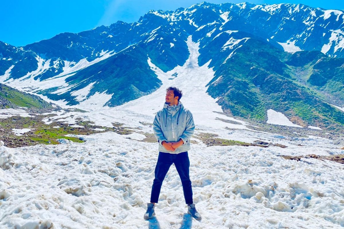 Traveler standing on a snowy mountain landscape with towering snow-capped peaks and a clear blue sky in the background.